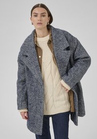 Žena oblečená v oversized kabáte s námornícko modrou a bielou vzorovanou farbou, pod ktorým má na sebe béžový sveter s copánkovým vzorom a hnedú košeľu, stojí proti šedému pozadiu.