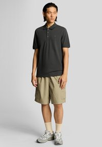 Polo nero con tre bottoni e un logo, abbinato a pantaloni corti beige. L'outfit è completato da calzini chiari e scarpe sportive.