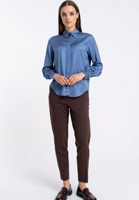 Kobieta stojąca z dłońmi złożonymi, ubrana w niebieską satynową bluzkę z detalami kołnierza, brązowe ciasne spodnie i ciemne mokasyny na białym tle.