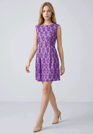 Joven mujer que lleva un vestido morado de encaje sin mangas y tacones altos beige, de pie contra un fondo gris claro y liso.