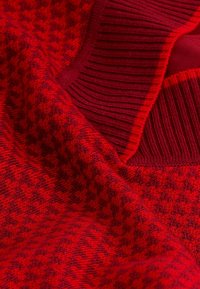 Nahaufnahme eines roten Strickpullovers mit geripptem Kragen und subtilem Hahnentrittmuster in dunkleren Rottönen.