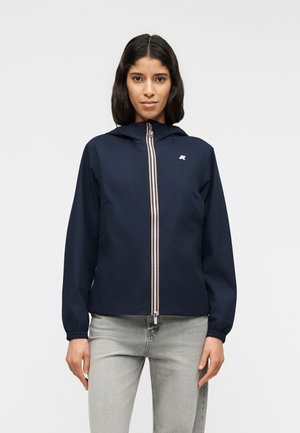 Žena s tmavými vlasmi po plecia, oblečená v tmavomodrej kapucňovej bunde s pruhovaným zipsom a svetlosivých džínsoch, na jednoliatom pozadí.