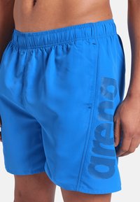 Torso maschile e parte superiore delle gambe che indossano pantaloni da bagno blu con vita elasticizzata e design di testo a pois neri sul lato.