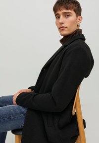 Cappotto di lana nera con un design strutturato. L'indossatore è seduto, abbinandolo a un dolcevita marrone a coste e jeans blu. Sedia di legno in vista.