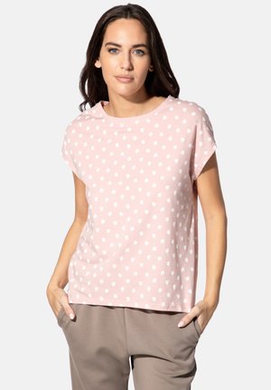 Frau mit dunklen Haaren, die ein rosa Kurzarmshirt mit weißen Punkten und taupefarbene Hose trägt, steht mit Händen in den Taschen vor weißem Hintergrund.