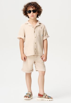 Beige korte mouwen shirt en bijpassende shorts van een gestructureerde stof, met een borstzak en bruine knopen. Gedragen met zwarte zonnebril en meerkleurige sandalen.