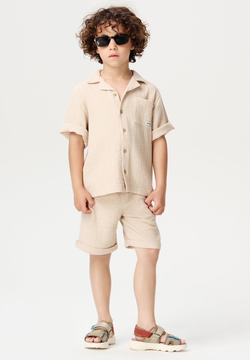 Beige korte mouwen shirt en bijpassende shorts van een gestructureerde stof, met een borstzak en bruine knopen. Gedragen met zwarte zonnebril en meerkleurige sandalen.