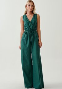 Jumpsuit verde senza maniche con scollatura a V profonda, vita cintrata e pantaloni a gamba larga. La texture morbida e il tessuto jersey creano una silhouette fluida.