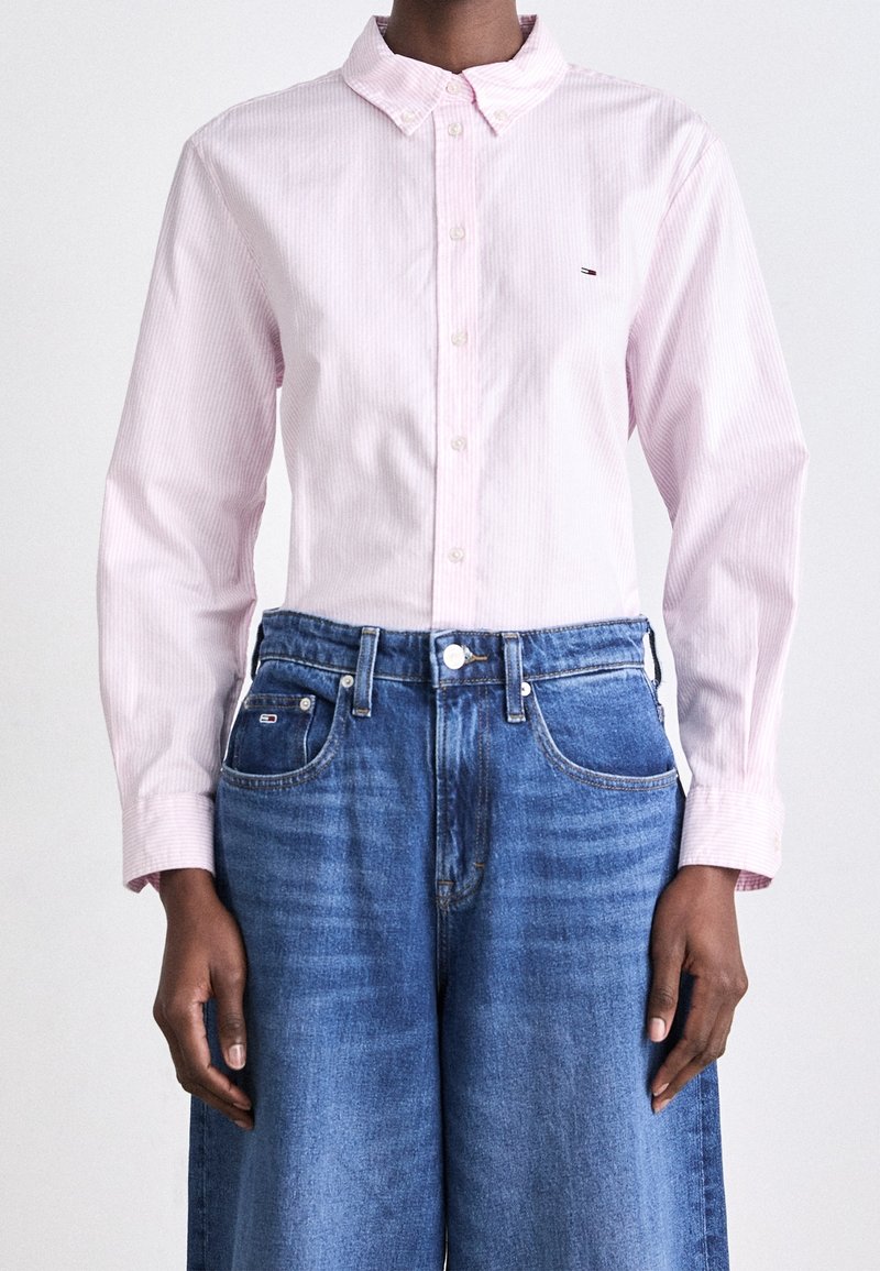 Personne portant une chemise rose clair à manches longues boutonnée et un jean bleu taille haute à jambes larges, debout devant un fond uni.
