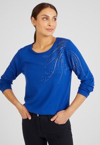 Blauwe trui met lange mouwen, ronde halslijn en versieringen met een patroon van strass steentjes op één schouder.