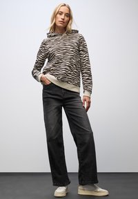 Bruine en crèmekleurige zebra patroonsweater met capuchon, gecombineerd met zwarte wijde jeans en beige sneakers, tegen een lichte achtergrond.