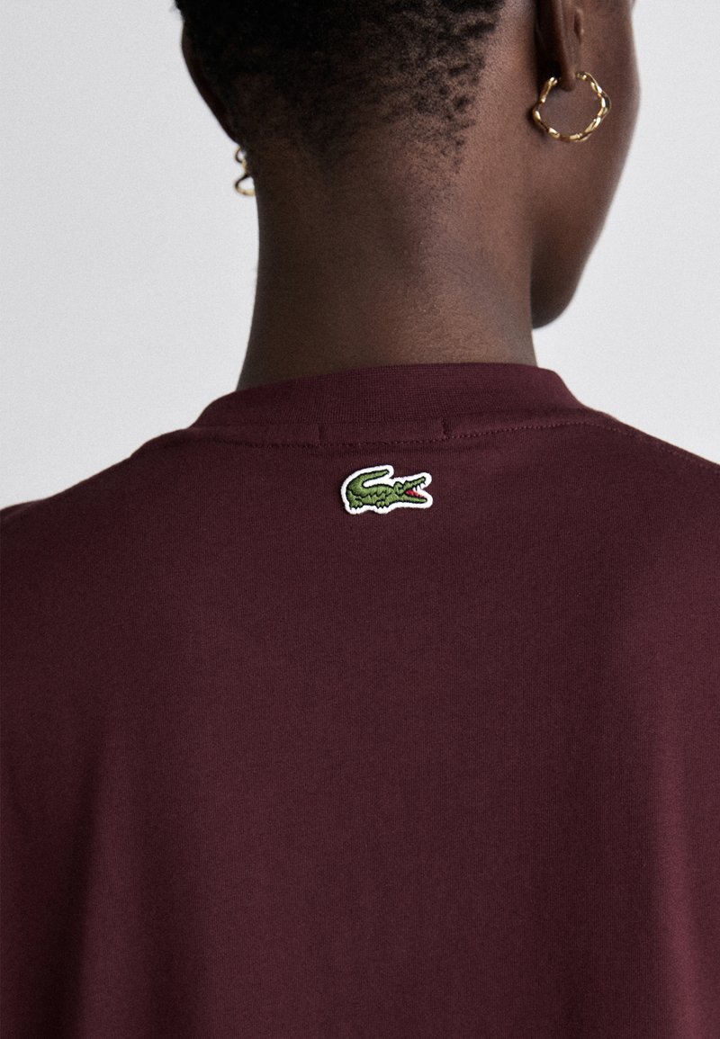 Camiseta de algodón burdeos con escote redondo y logo de cocodrilo verde bordado en la parte posterior. Textura suave y diseño sencillo.