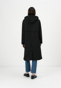 Casaco de chuva preto com capuz e corte relaxado, apresentando mangas longas e uma abertura nas costas. Combinado com jeans curtos azuis e sapatos pretos.