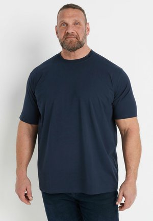 Brodaty mężczyzna z krótkimi włosami, ubrany w prostą, ciemnogranatową koszulkę z krótkim rękawem, stojący na białym tle.