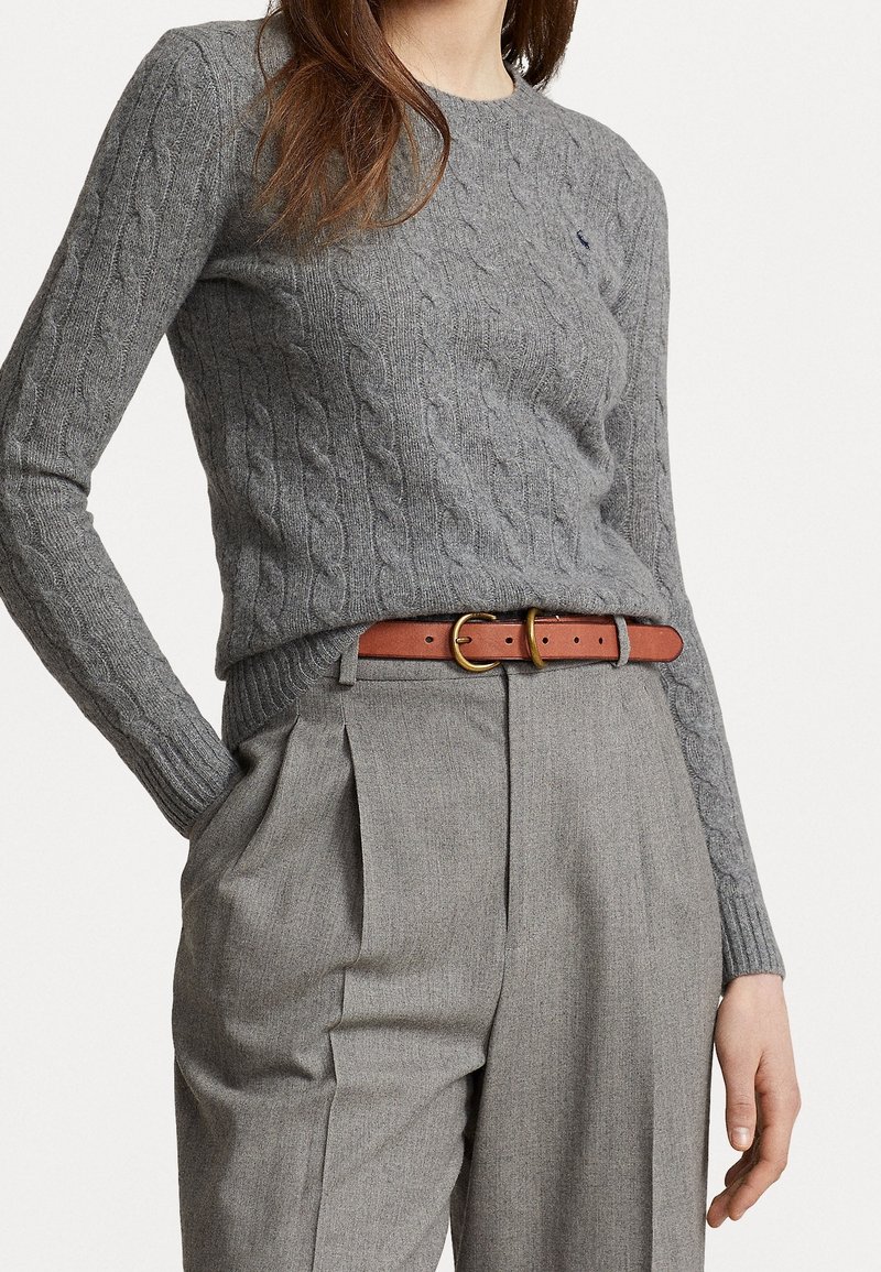 Femme portant un pull gris en maille torsadée, rentré dans un pantalon plissé gris taille haute avec une ceinture en cuir marron à boucles dorées.