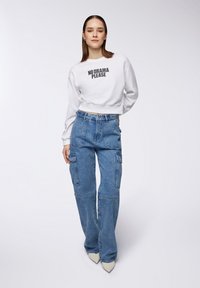 Felpa bianca a maniche lunghe con testo nero "NO DRAMA PLEASE" abbinata a pantaloni cargo in denim blu dal fit ampio, dotati di tasche laterali e vestibilità rilassata.