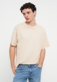 Μπεζ βαμβακερό T-shirt με χαλαρή εφαρμογή, κοντά μανίκια και στρογγυλή λαιμόκοψη. Διακριτική λεπτομέρεια με λογότυπο στο στήθος, συνδυασμένο με ανοιχτόχρωμο μπλε τζιν.