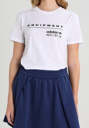 Frau trägt ein weißes Adidas Equipment T-Shirt mit schwarzem Text und einen marineblauen Rock mit elastischem Bund.