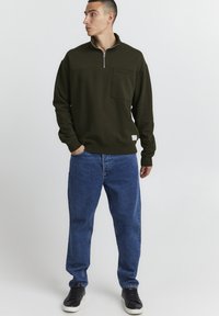 Mørkegrøn fleece sweatshirt med lynlås og en frontlomme, kombineret med blå denim jeans og sorte sneakers. Enkel design, blød tekstur.