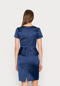 Marinblå satindress med korta ärmar, peplummidja och en slits bak. Har en dragkedja bak och en mjuk, glänsande yta.