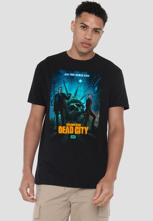 Junger Mann trägt bei Nacht ein schwarzes T-Shirt mit dem Aufdruck "Walking Dead Dead City", darauf zwei Charaktere und ein verfallener Kopf der Freiheitsstatue.