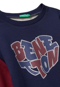 Felpa blu navy con dettagli color bordeaux. Presenta un grande logo "BENETTON" multicolore in glitter argento. Collo e polsini a costine.