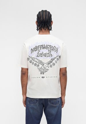 Muškarac s pletenicama nosi bijelu majicu s tekstom u plamenu "Huntington Beach", grafikom orla i natpisom "Design Studios" na leđima.