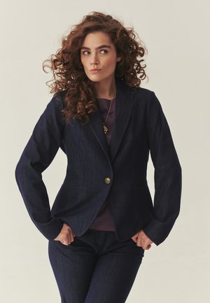 Vrouw met krullend haar, gekleed in een donkerblauw pak en paarse top, staand met handen in de zij, kijkt opzij tegen een neutrale achtergrond.