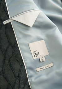Jasnoniebieska satynowa tkanina z białym kontrastującym kołnierzykiem, nałożona na teksturowaną czarną koronkę. Metki mówią "NEXT EDIT." oraz "REGULAR FIT."