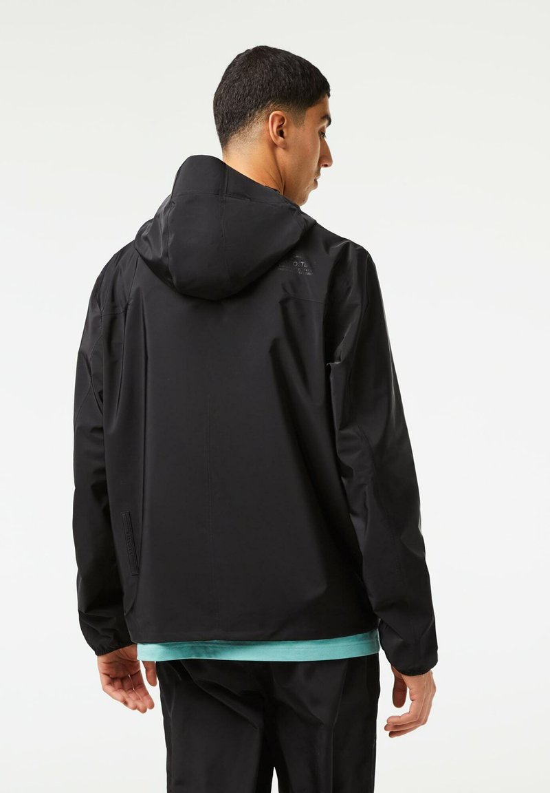 Veste noire avec capuche, dotée d'une texture lisse et d'un design minimal. Portée sur une chemise turquoise, avec des poches latérales et une coupe décontractée.