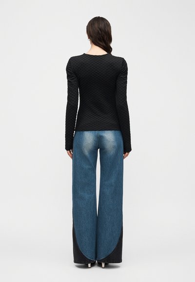 Γυναίκα που φοράει μαύρο μπλουζάκι με υφή και μακριά μανίκια και μπλε τζιν φαρδιάς γραμμής με σκούρες καμπυλωτές λεπτομέρειες, στέκεται πλάτη σε λευκό φόντο.