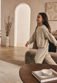 Set di abbigliamento comodo color beige chiaro con texture a coste; maglia a maniche lunghe e pantaloni abbinati. Tessuto morbido, vestibilità rilassata e maniche ampie e fluide.