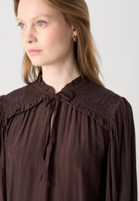 Brun, flydendebluse lavet af letvægtsstof med smocked detaljer ved kraven og en snøring. Blødt, tekstureret finish.