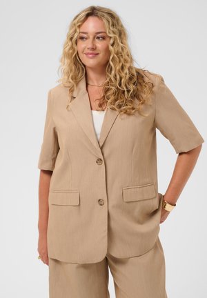 Vrouw met krullend blond haar, gekleed in een beige kort mouwen blazer en bijpassende broek, staand met één hand in de zak, zacht glimlachend.