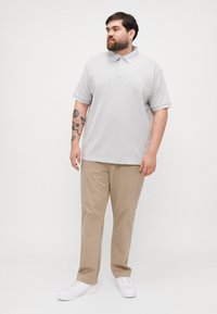 Svetlo siva polo majica s kratkimi rokavi, ohlapen kroj in diskreten logotip. V kombinaciji s bež hlačami in belimi supergami, kar ustvarja sproščen videz.