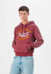 Sudadera color burdeos con cordones, bolsillo frontal y texto impreso en blanco y amarillo. Combinada con jeans azul claro para un look casual.