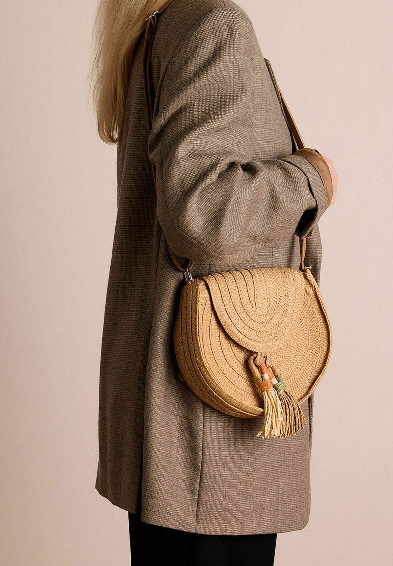 Borsa a tracolla di paglia intrecciata rotonda in beige con una superficie testurizzata, dotata di due frange verdi e arancioni. Tracolla regolabile.