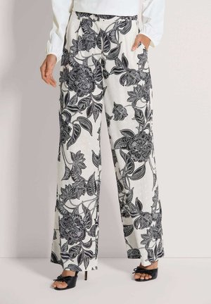 Weiße Hosen mit weitem Bein und schwarzem Blumenmuster, kombiniert mit schwarzen Sandalen mit Absatz und einem weißen Langarmshirt, die Hände ruhen seitlich.