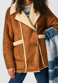 Brun faux shearling-jacka med stor krämfärgad krage, framknappar och fickor med lappar. Har en mjuk textur och en avslappnad silhuett.