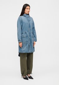 Μακρύ τζιν παλτό σε ανοιχτό μπλε χρώμα με κουκούλα, κουμπιά μπροστά, δύο τσέπες στη μέση και κορδόνι στο τελείωμα. Συνδυασμένο με ελαιοπράσινο παντελόνι.