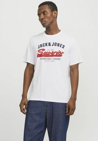 White cotton t-shirt with printed logos in navy and red, featuring short sleeves and a loose fit. Worn with dark denim trousers.