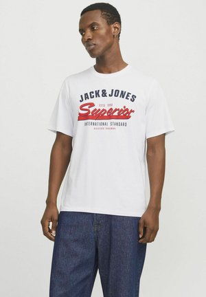 Witte katoenen t-shirt met bedrukte logo's in marineblauw en rood, met korte mouwen en een losse pasvorm. G gedragen met donkere spijkerbroek.
