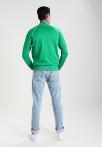 Felpa sportiva verde con dettagli bianchi, orlo a coste e maniche lunghe; abbinata a jeans azzurri chiari e sneaker bianche.