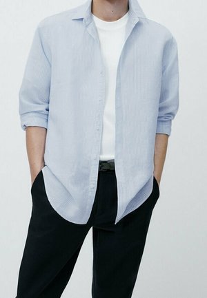 Person trägt ein hellblau gestreiftes Hemd über einem weißen T-Shirt, schwarze Hose und hat die Hände in den Taschen.