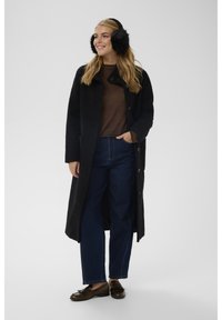 Femme portant un long manteau noir texturé, un pull marron, un jean bleu foncé, des mocassins marron et des cache-oreilles noirs en fourrure, debout avec une main dans la poche.