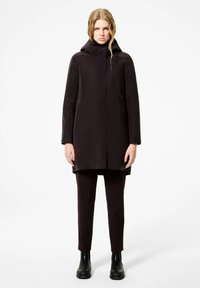 Cappotto impermeabile nero con cappuccio, caratterizzato da una vestibilità dritta e tasche laterali, abbinato a pantaloni neri aderenti e stivali in gomma alla caviglia.