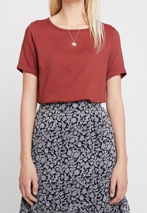 Kobieta w rdzawym, krótki rękaw topie i granatowej, wysokiej talii spódnicy z białym kwiatowym wzorem.