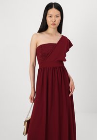 Rochie bordeaux cu o singură umăr, având un corset din dantelă, fustă vaporoasă și un accent cu volane. Purtată cu o geantă mică aurie.