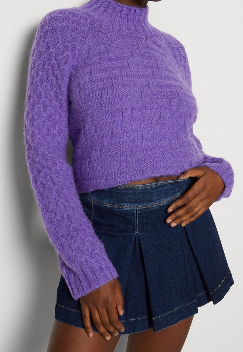 Femme porte un pull en maille violet duveteux à col haut et une jupe en jean plissée bleu foncé.