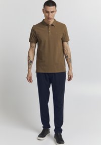 Brun polotröja med krage, korta ärmar och tre knappar. Bärs med marinblå byxor och svarta sneakers. Tatueringar synliga på armarna.